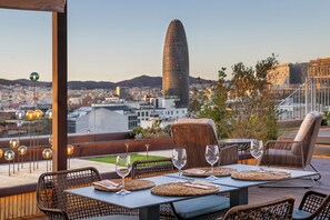 Un espacio exterior para comer con mobiliario de mimbre y una mesa dispuesta para dos, con vistas a un paisaje urbano que destaca una torre singular.