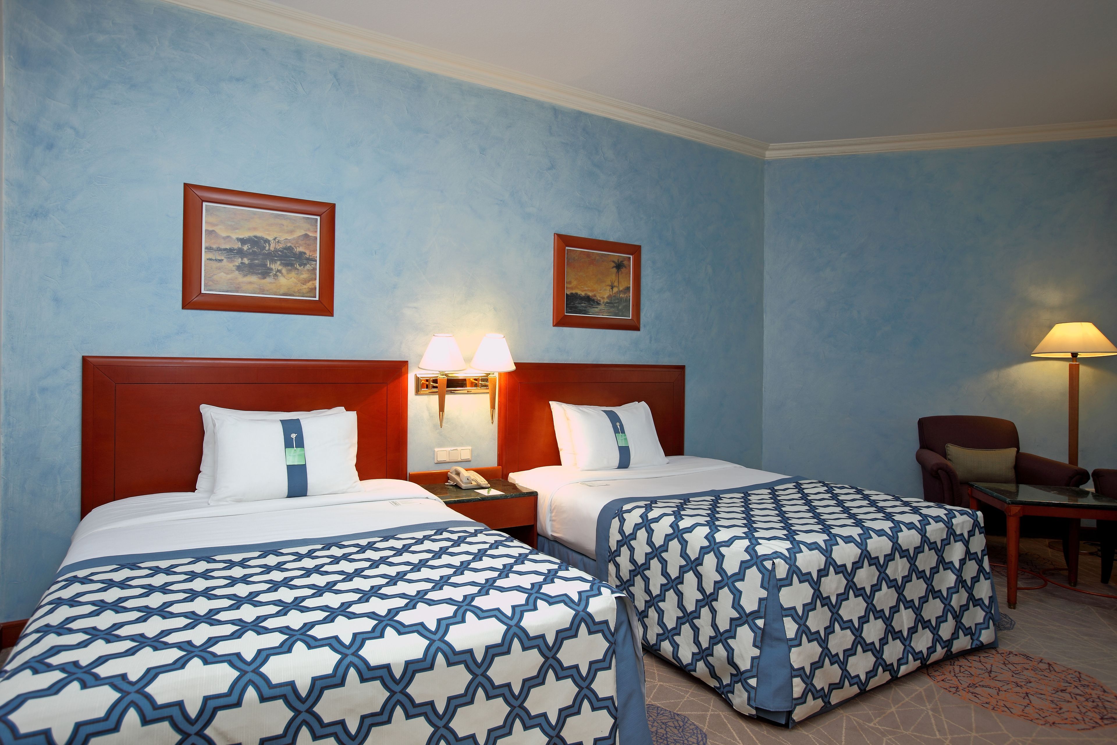 A hotel room with two beds, a nightstand with a lamp, a chair, and two framed pictures on the wall.