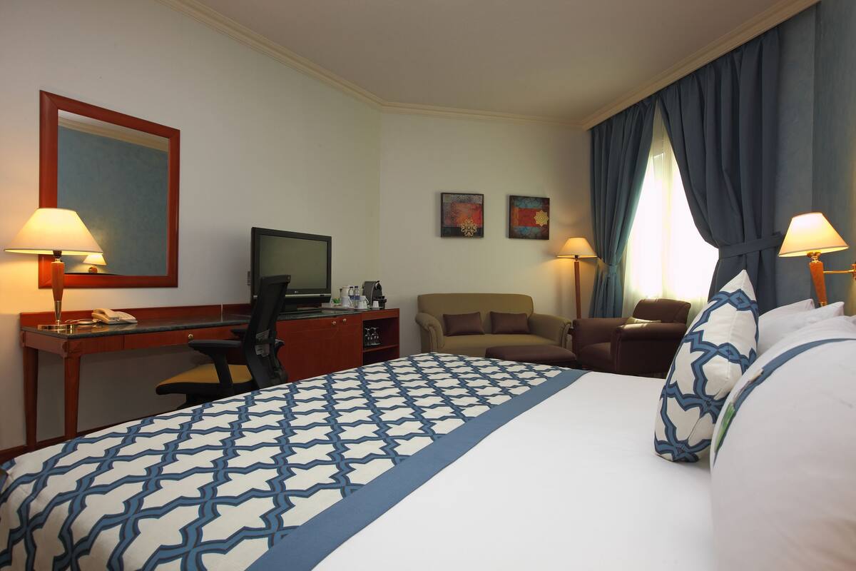 A hotel room with a large bed, a desk with a computer, a chair, and two wall-mounted lamps.
