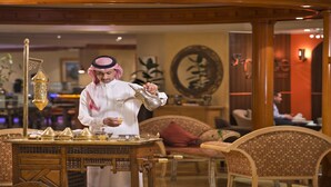 A traditional setting with a person pouring a drink, a decorative lamp, and a table with various items.