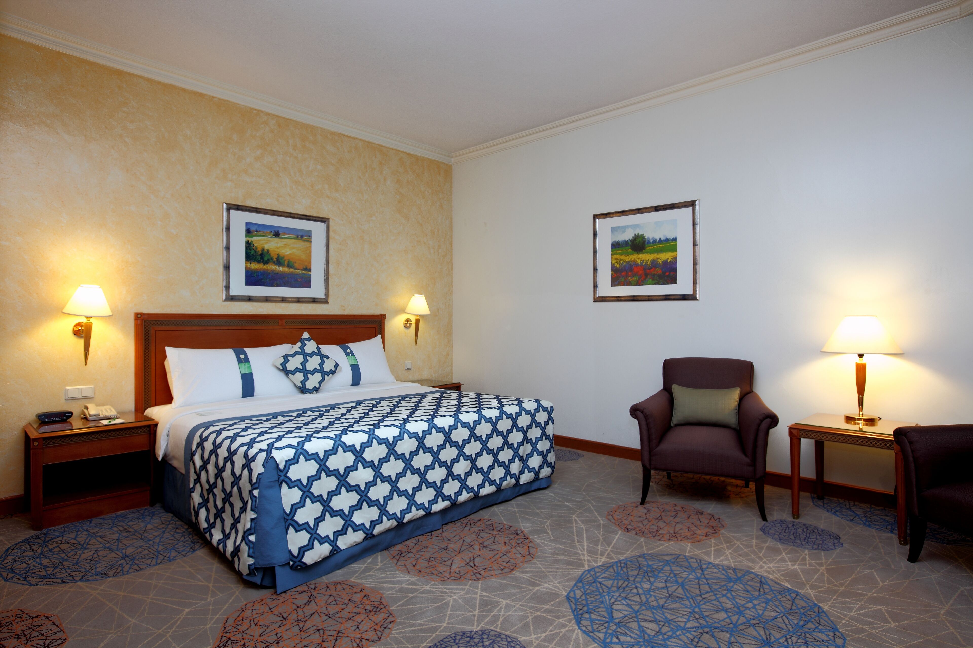 A hotel room with a large bed, two bedside tables with lamps, a chair, and two framed pictures on the walls.