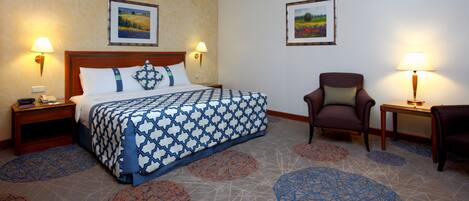 A hotel room with a large bed, two bedside tables with lamps, a chair, and two framed pictures on the walls.