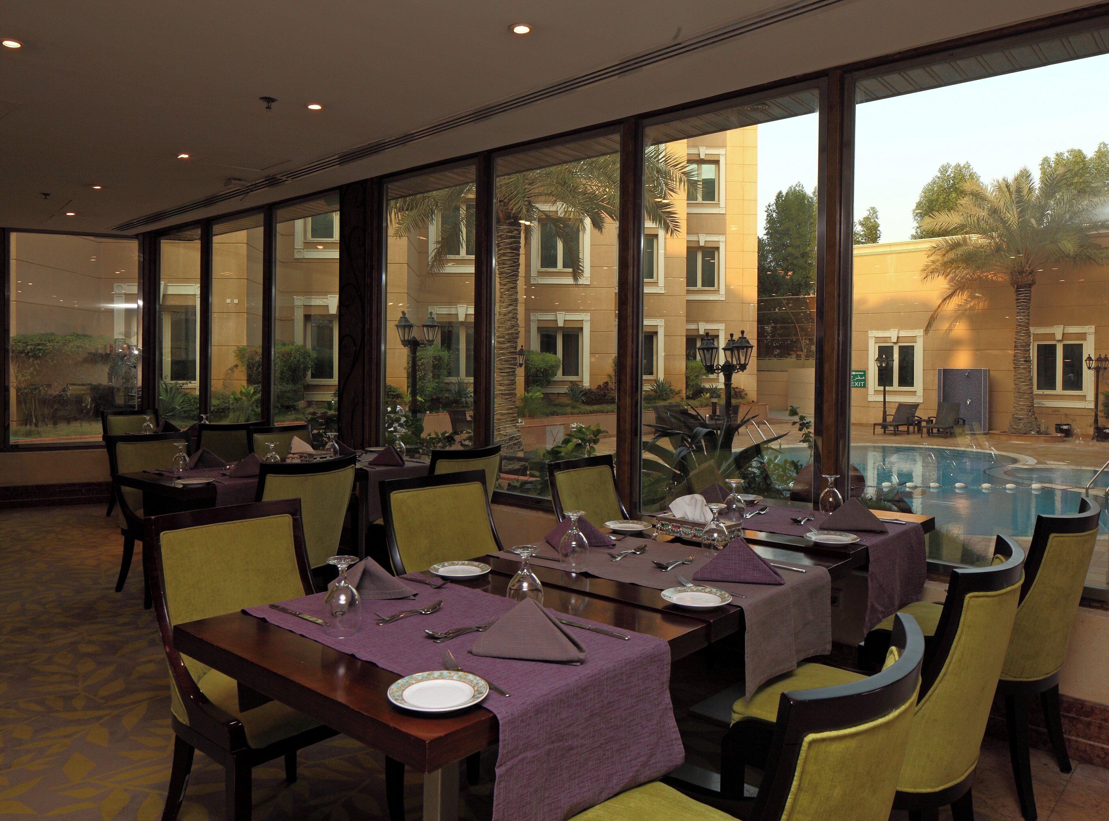 Un comedor con mesas dispuestas para el servicio, amplias ventanas que ofrecen vistas a una piscina y un área exterior, y piso alfombrado.