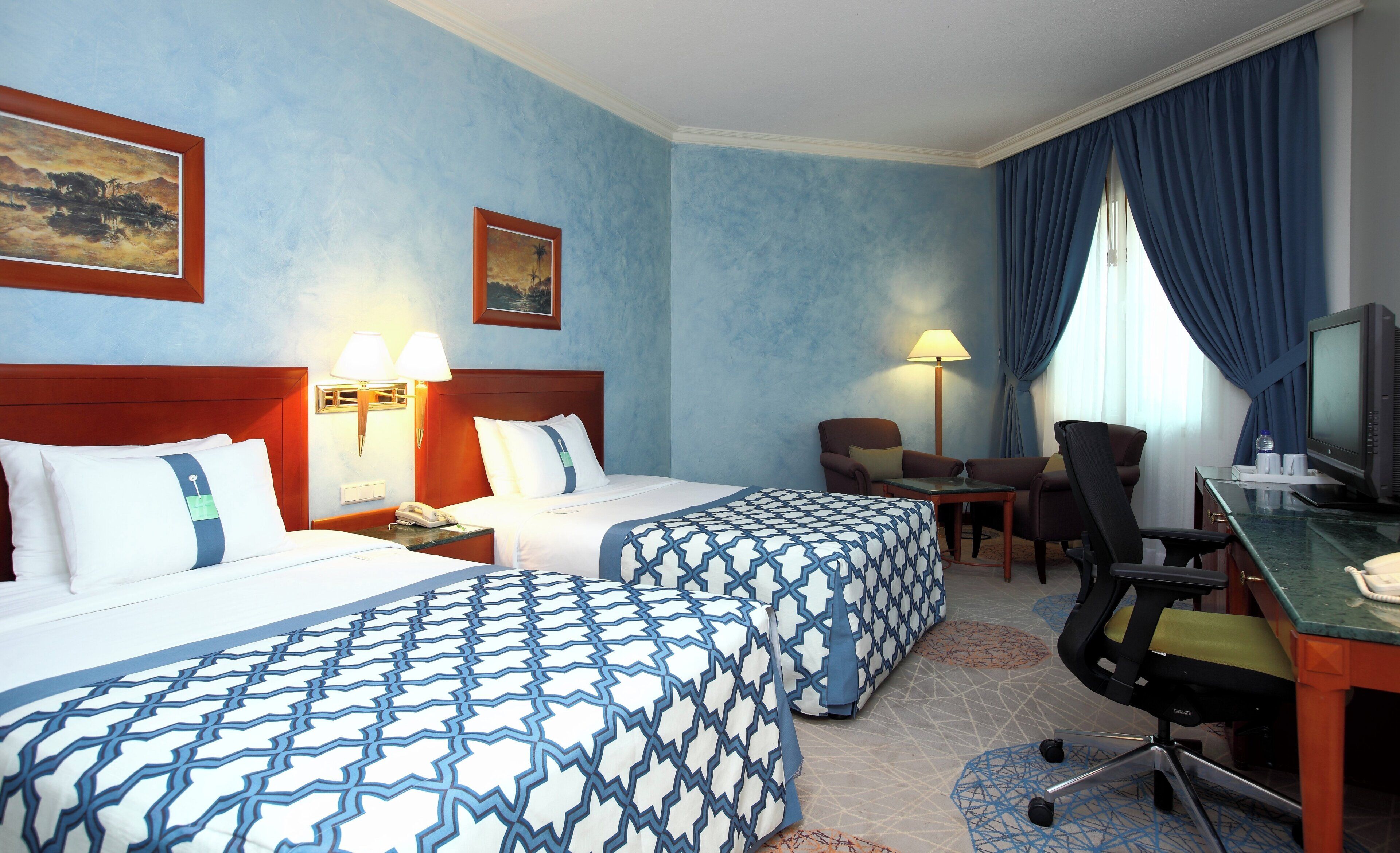 Habitación de hotel con dos camas, un escritorio con computadora, una silla, una lámpara y un cuadro en la pared.