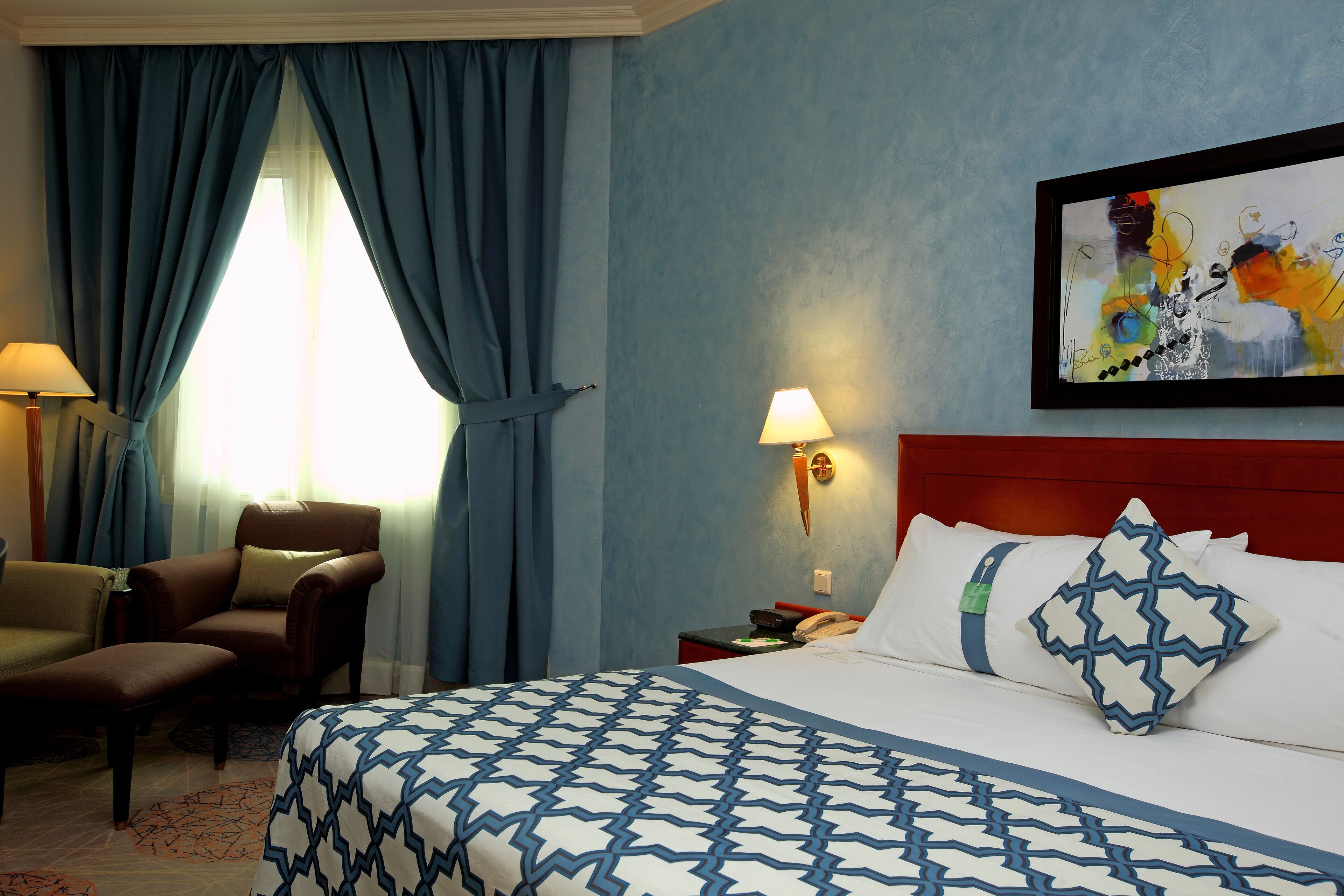 A hotel room with a bed, a chair, a nightstand, and a painting on the wall.