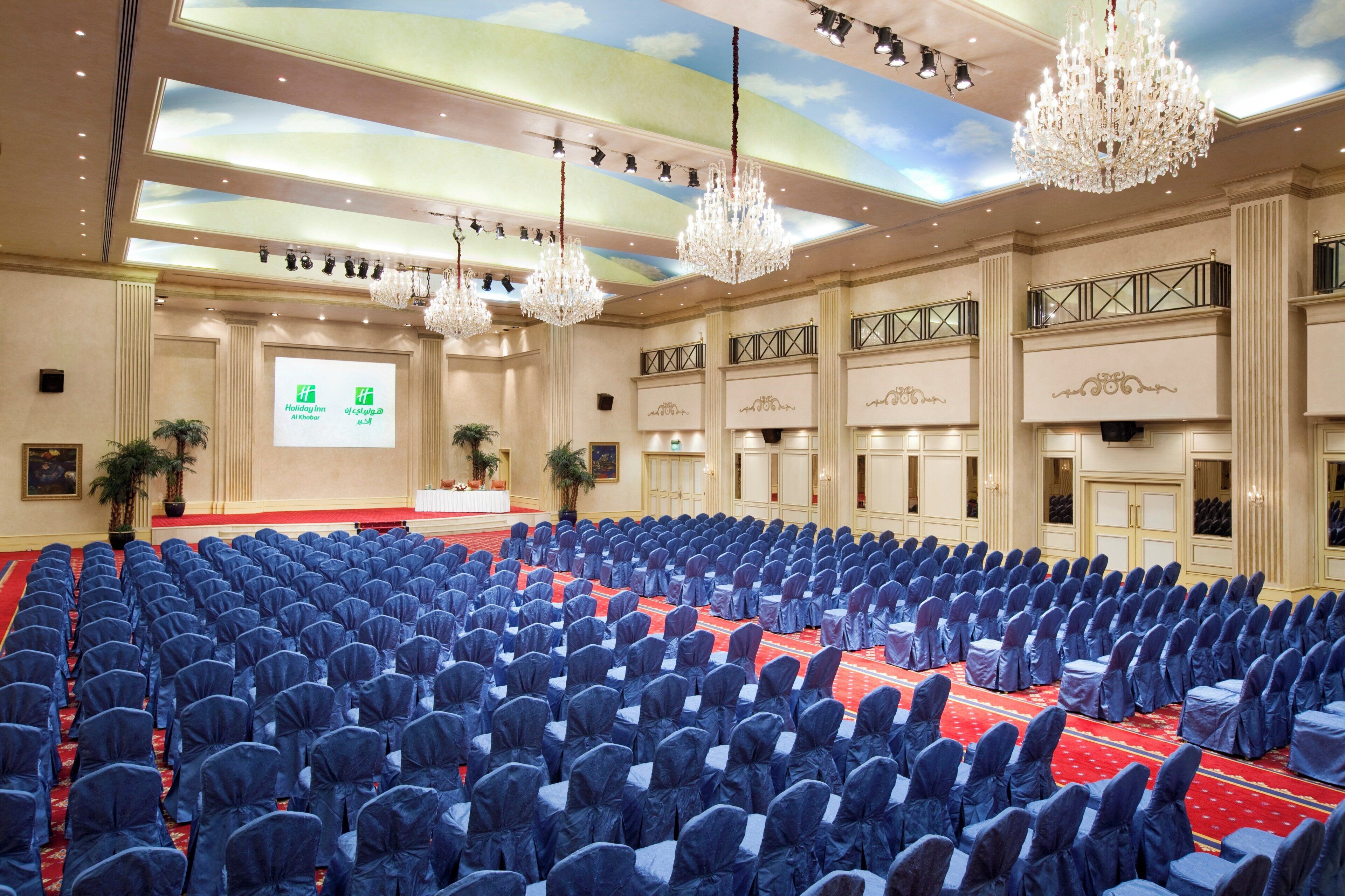 A large conference room with rows of chairs, a stage with a presentation screen, chandeliers, and a red carpet.