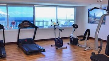 Ein Fitnessraum mit Laufbändern, Crosstrainern und Krafttrainingsgeräten, mit Blick auf das Meer durch große Fenster.