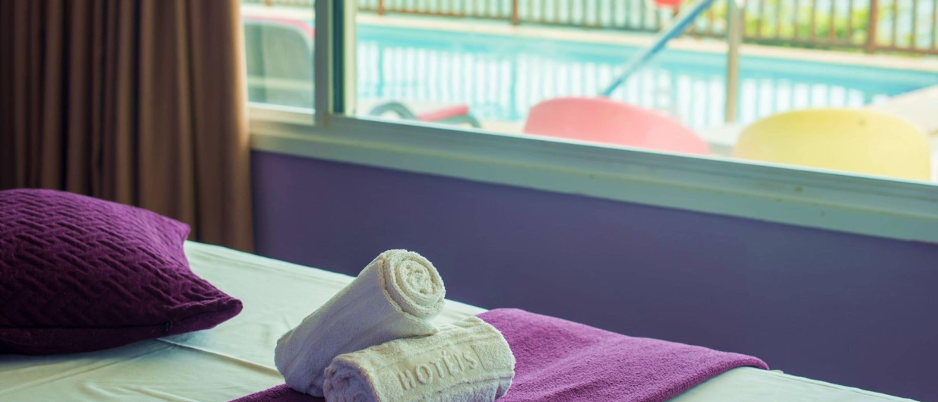 A bed with a purple towel and a rolled towel with "HOTELS" embossed on it.
