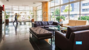 Um hall de entrada com sofá, banquinho e mesa de centro, amplas janelas e uma pessoa caminhando.