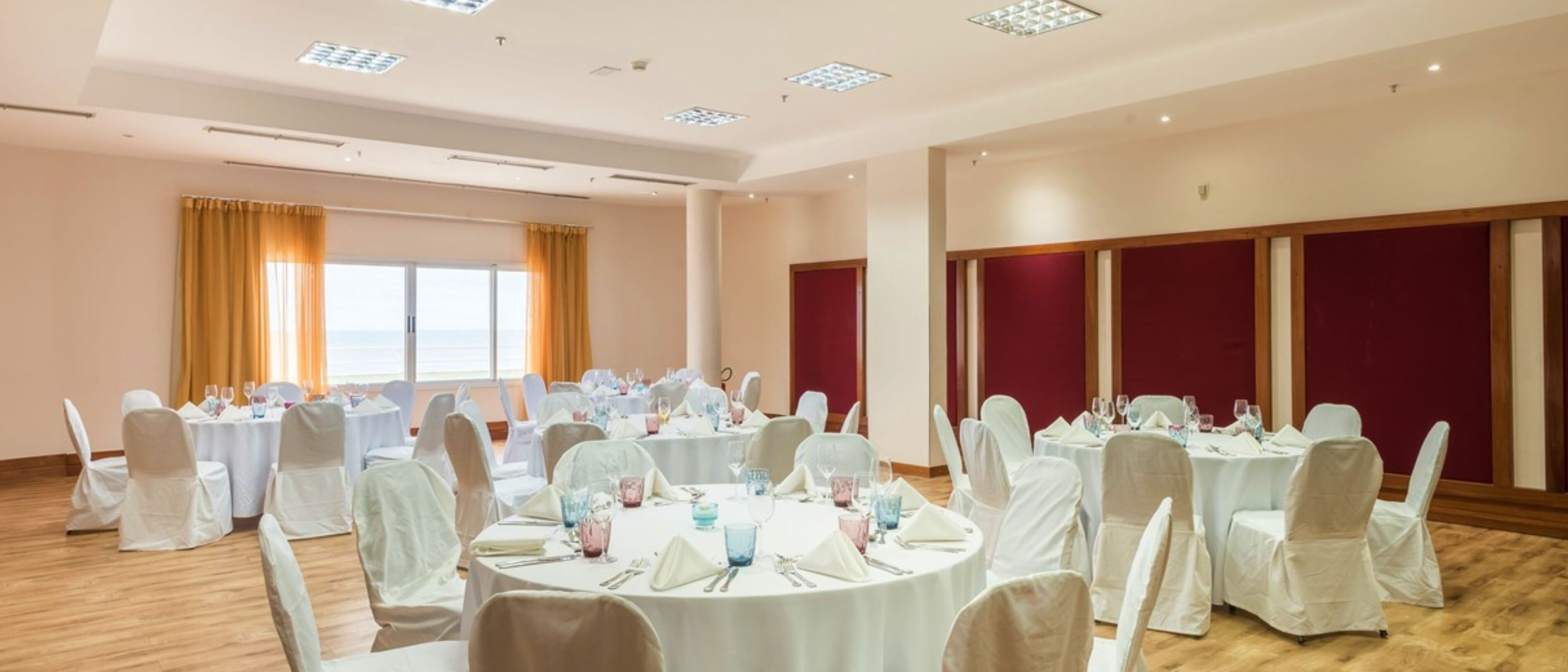 A banquet hall with round tables set for dining, white chairs, and a view of the sea through large windows.