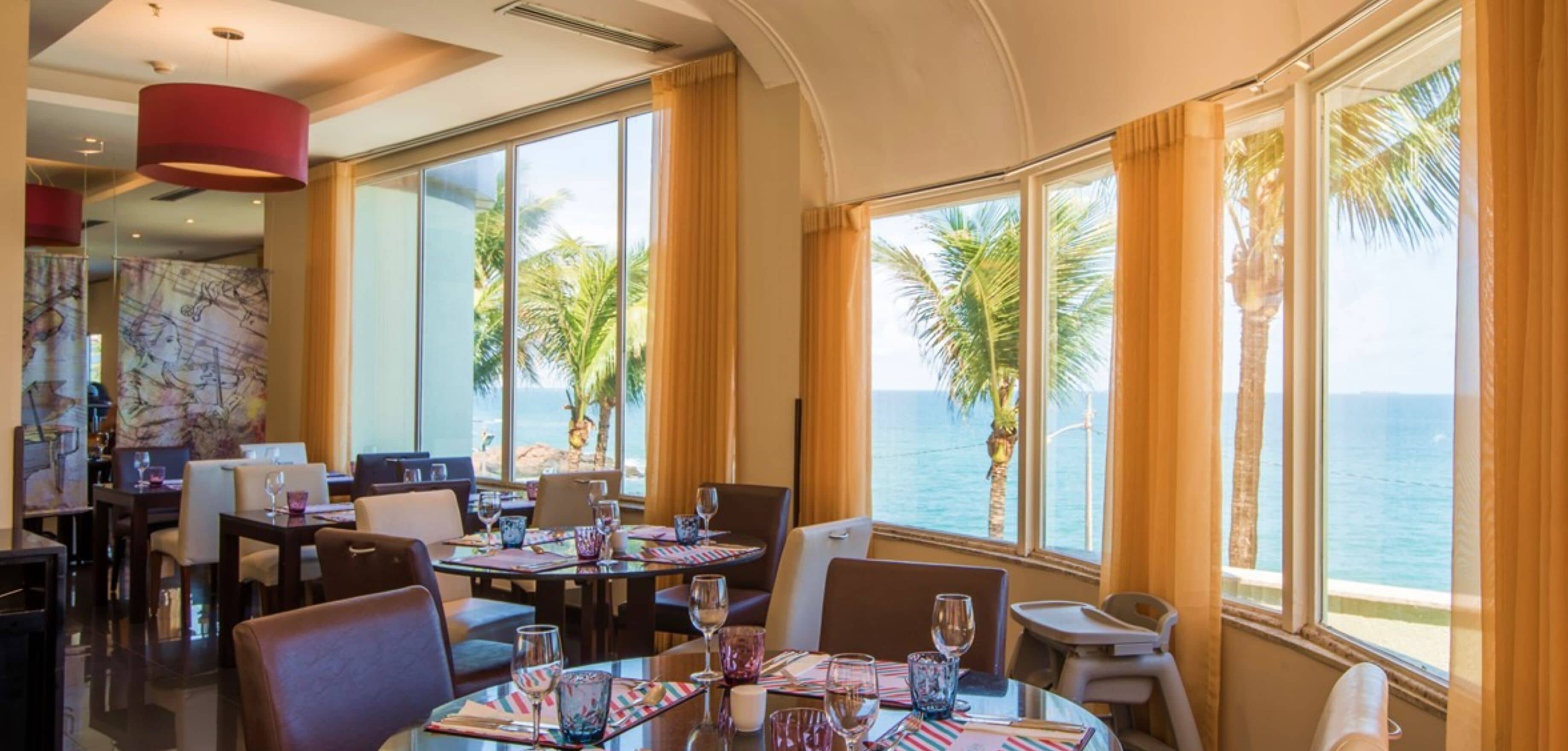 A restaurant with tables set for dining, large windows with a view of the sea, and tropical plants outside.