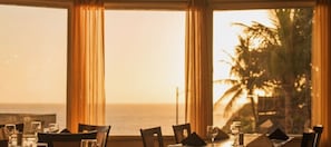 A dining area with tables and chairs, large windows with curtains, and a view of the ocean.
