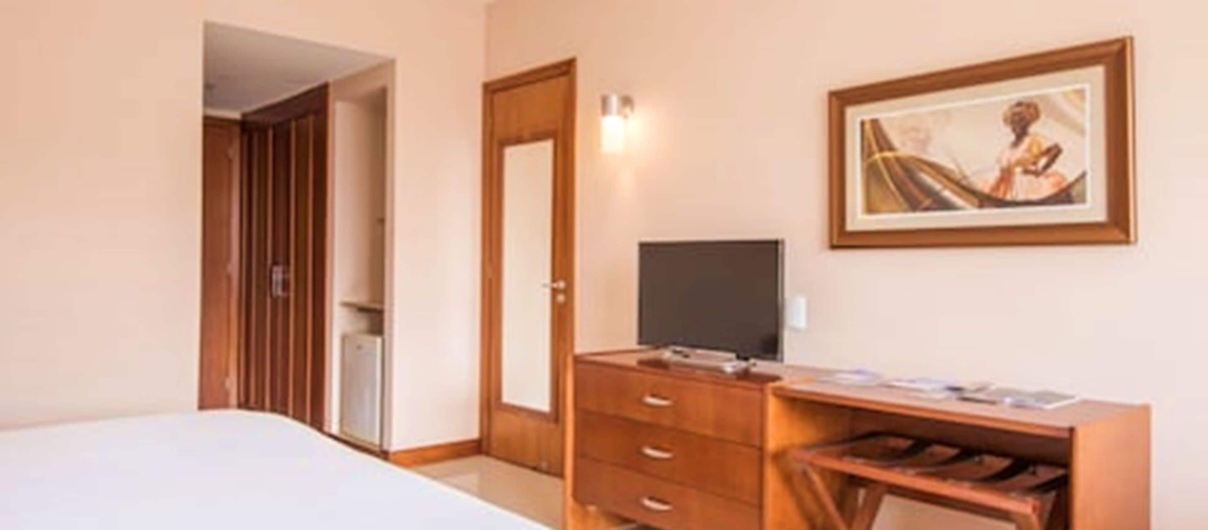 Une chambre d’hôtel avec un lit, un bureau équipé d’un ordinateur, une télévision et un tableau encadré au mur.