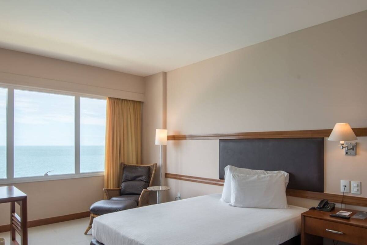 Ein Hotelzimmer mit Bett, Nachttisch, Lampe, Stuhl und Meerblick durch große Fenster.