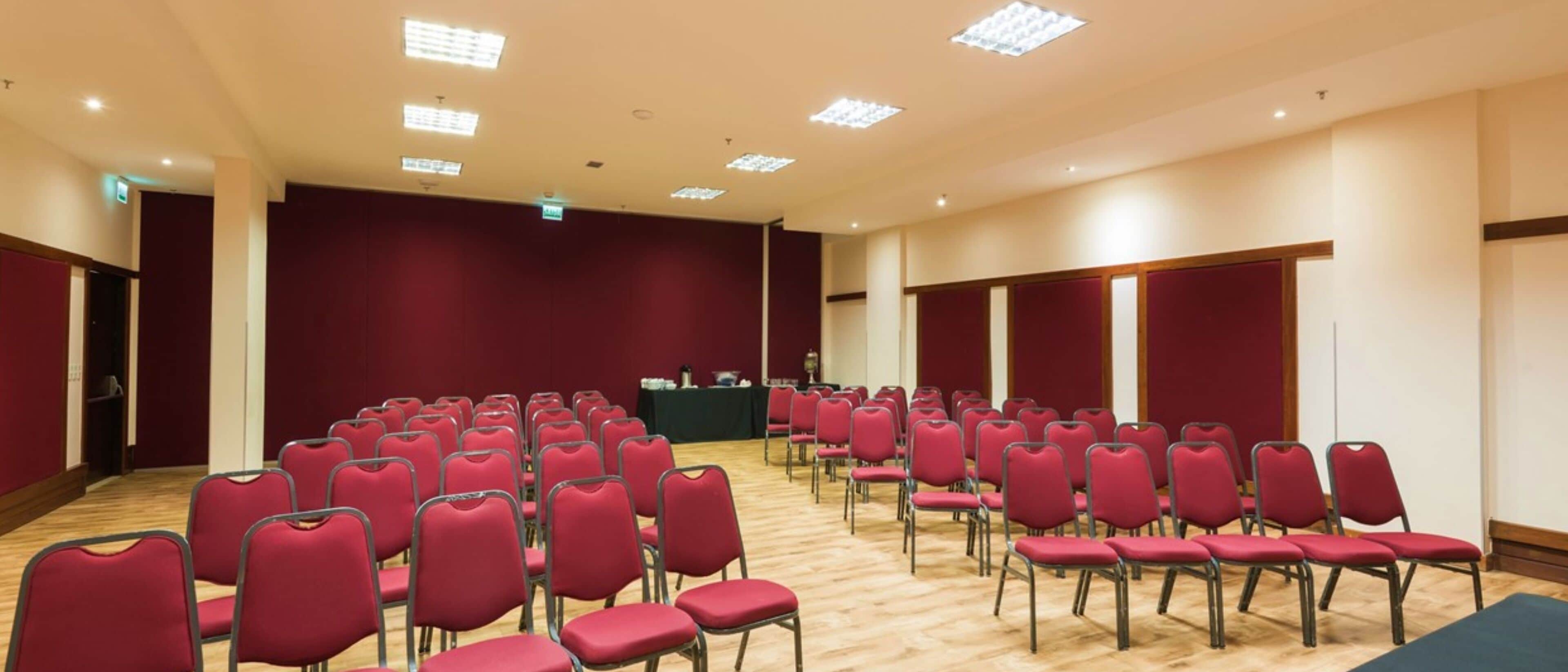 Uma sala de conferência com fileiras de cadeiras vermelhas, um palco com um pódio e uma mesa com toalha verde.