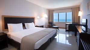 Quarto de hotel com uma cama grande, uma escrivaninha com um computador, uma cadeira e vista para o mar pela janela.