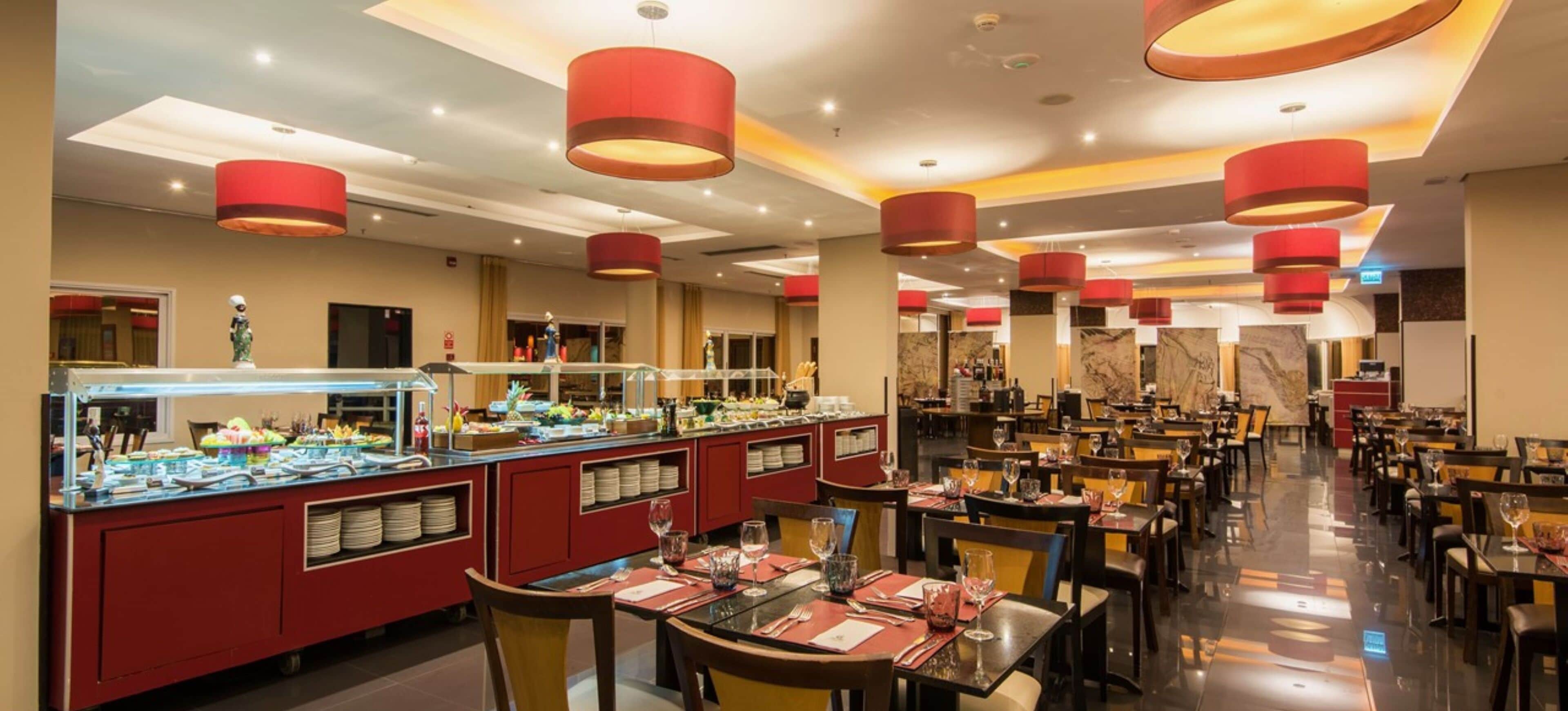 A hotel dining area with multiple tables and chairs, a buffet with various food items, and red cylindrical light fixtures on the ceiling.