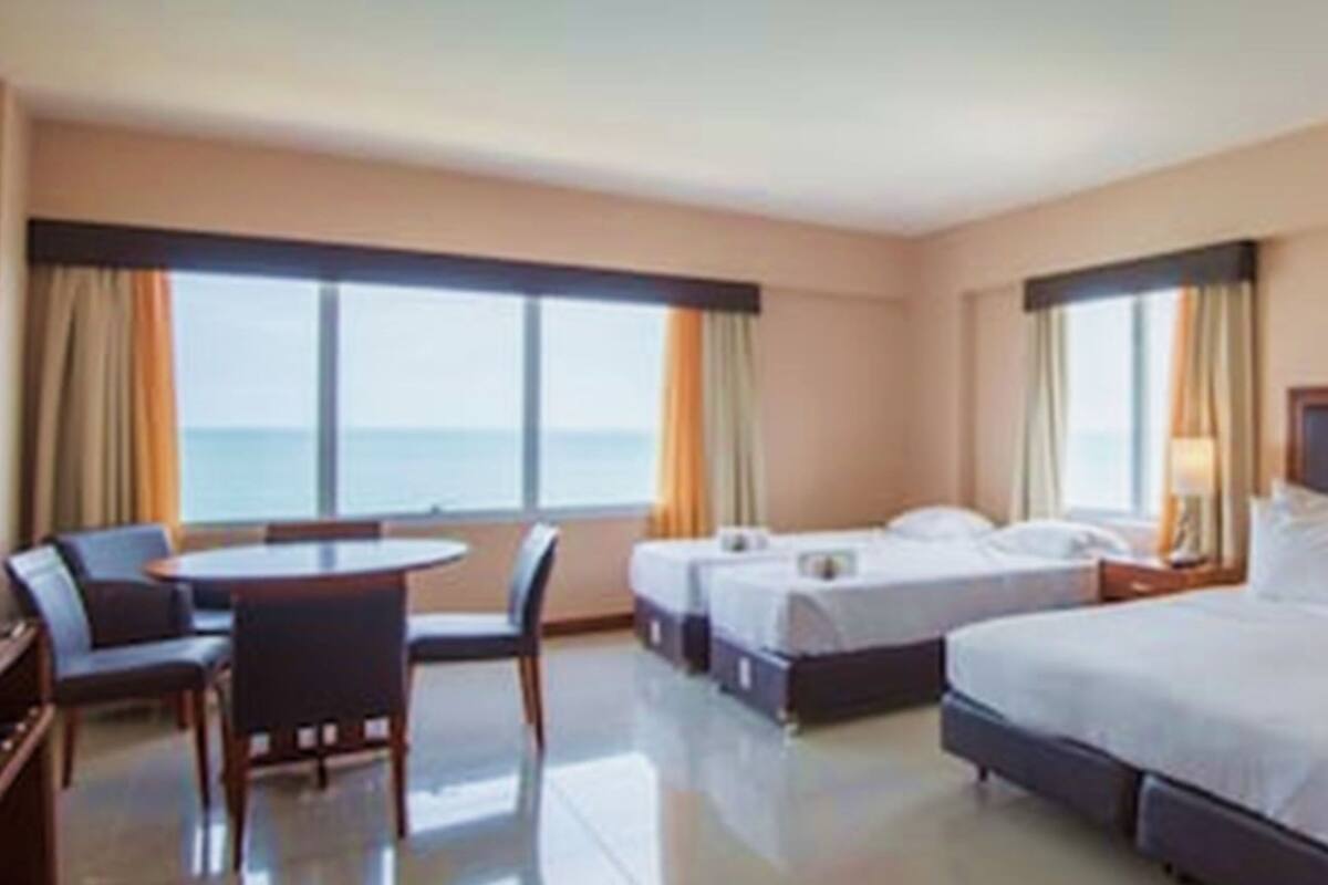 Ein Hotelzimmer mit einem großen Bett, zwei Einzelbetten, einem Schreibtisch und einem Sessel. Große Fenster mit Vorhängen und eine Lampe auf dem Nachttisch.