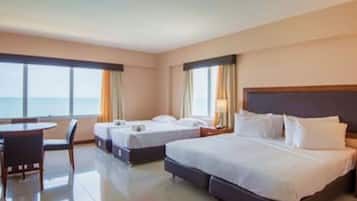Une chambre d’hôtel avec un grand lit, deux petits lits, un bureau et une chaise. De grandes fenêtres sont présentes, avec des rideaux, et une lampe se trouve sur la table de chevet.