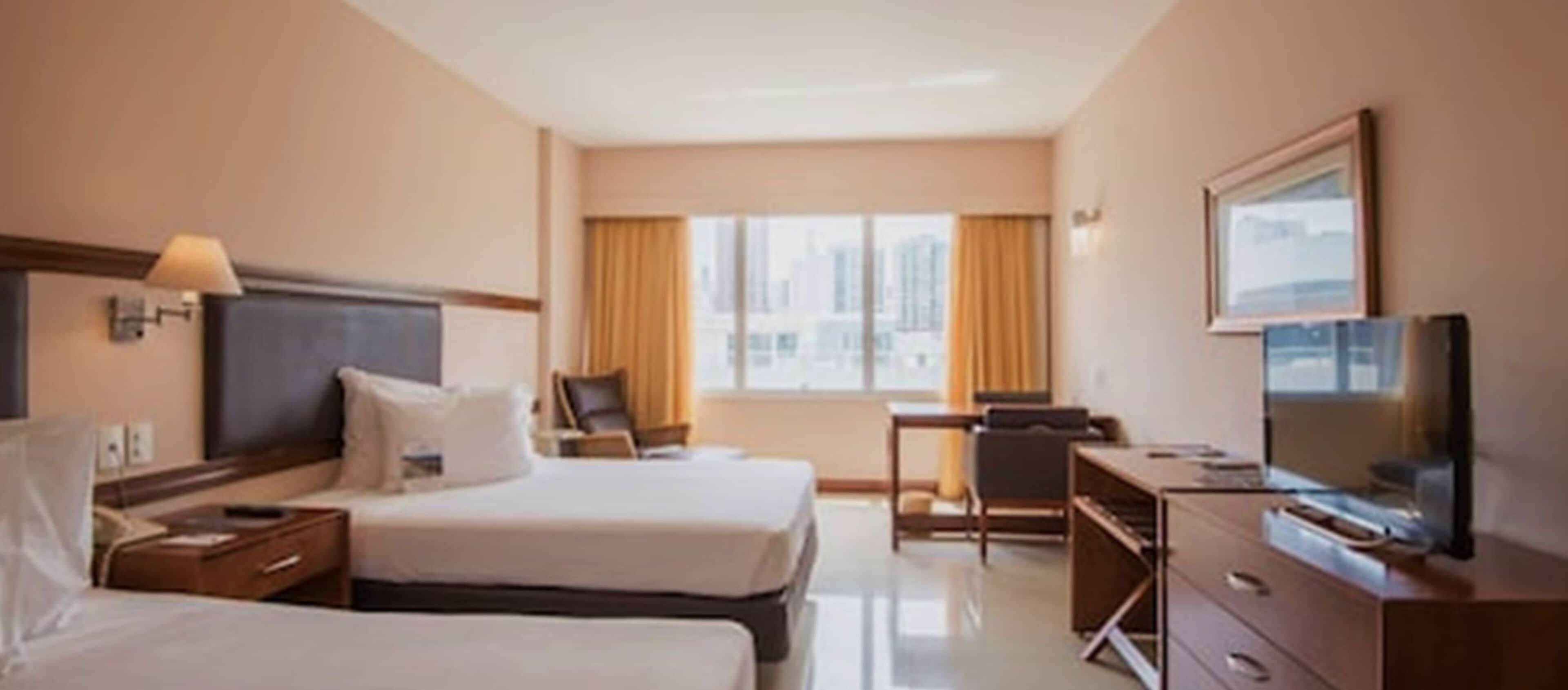 Une chambre d’hôtel avec deux lits, un bureau, une chaise, une télévision et une fenêtre avec des rideaux.
