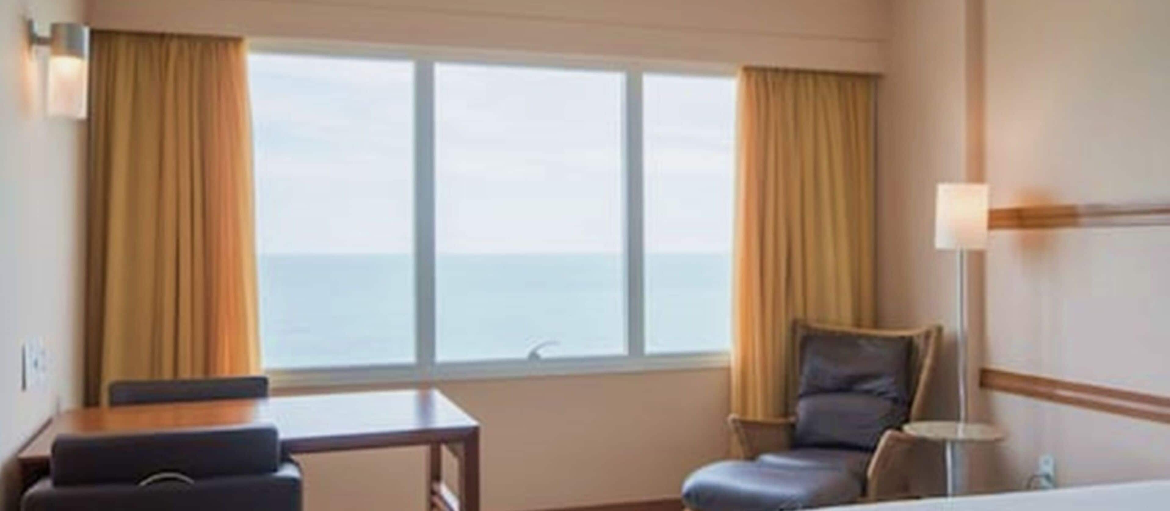 A hotel room with a large window, a desk, a chair, and a lamp.