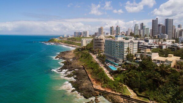 Un paisaje urbano costero con un hotel frente a la playa que cuenta con piscina, rodeado de edificios altos y un cielo azul despejado.
