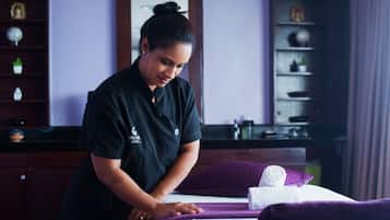 Uma pessoa de uniforme preto atendendo um hóspede em um quarto com colcha roxa, uma toalha enrolada e uma estante com diversos objetos.