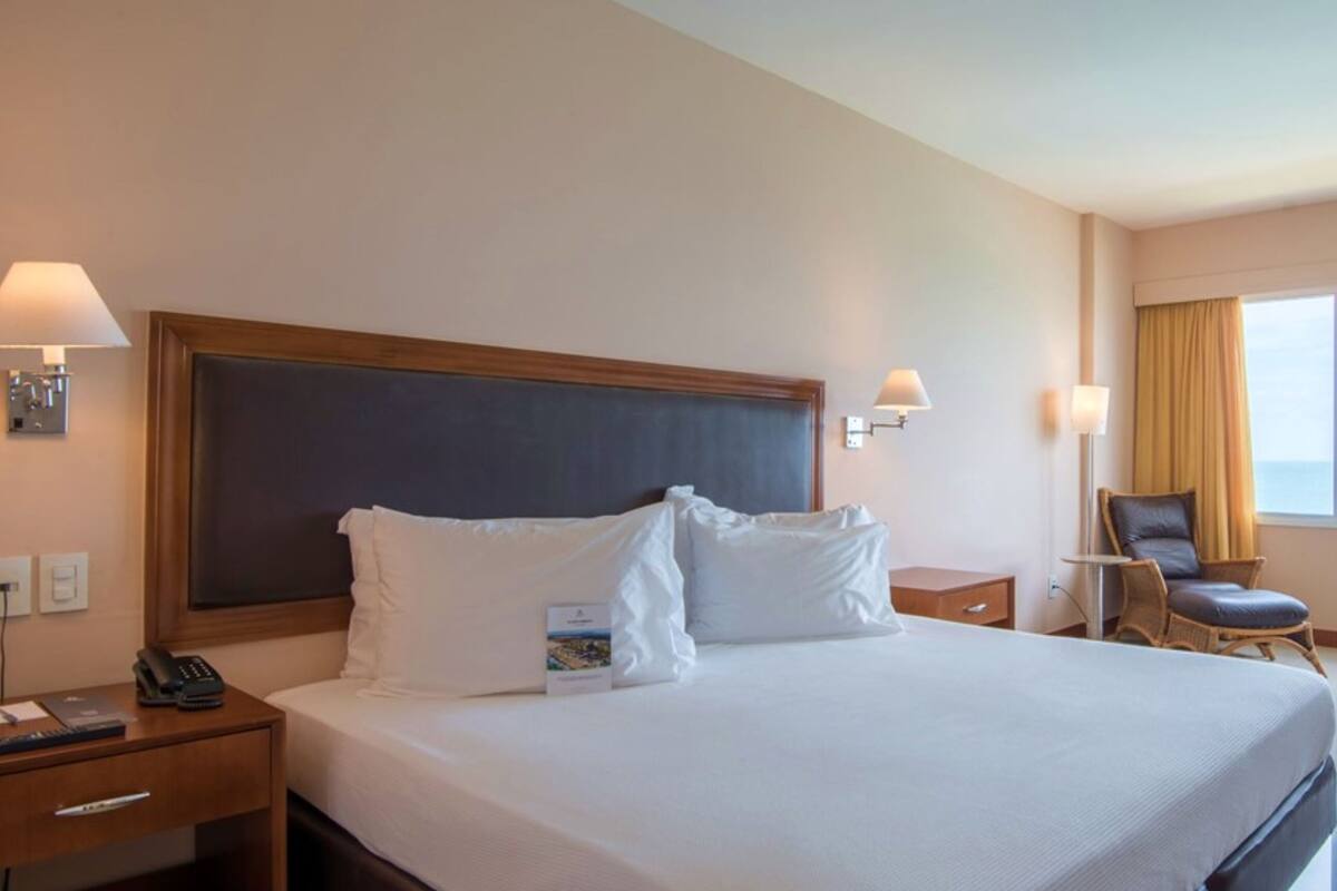 Ein Hotelzimmer mit einem großen Bett, einem Schreibtisch mit Stuhl, einer Lampe und Meerblick.