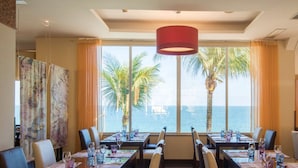 Uma área de refeições com mesas preparadas para quatro pessoas, grandes janelas com vista para palmeiras e o mar, e uma luminária pendente vermelha sobre as mesas.