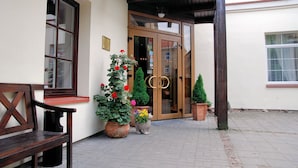 A wooden bench, a potted plant with red flowers, and a building entrance with a glass door and a wooden frame. - City Gate Hotel (Vilnius)