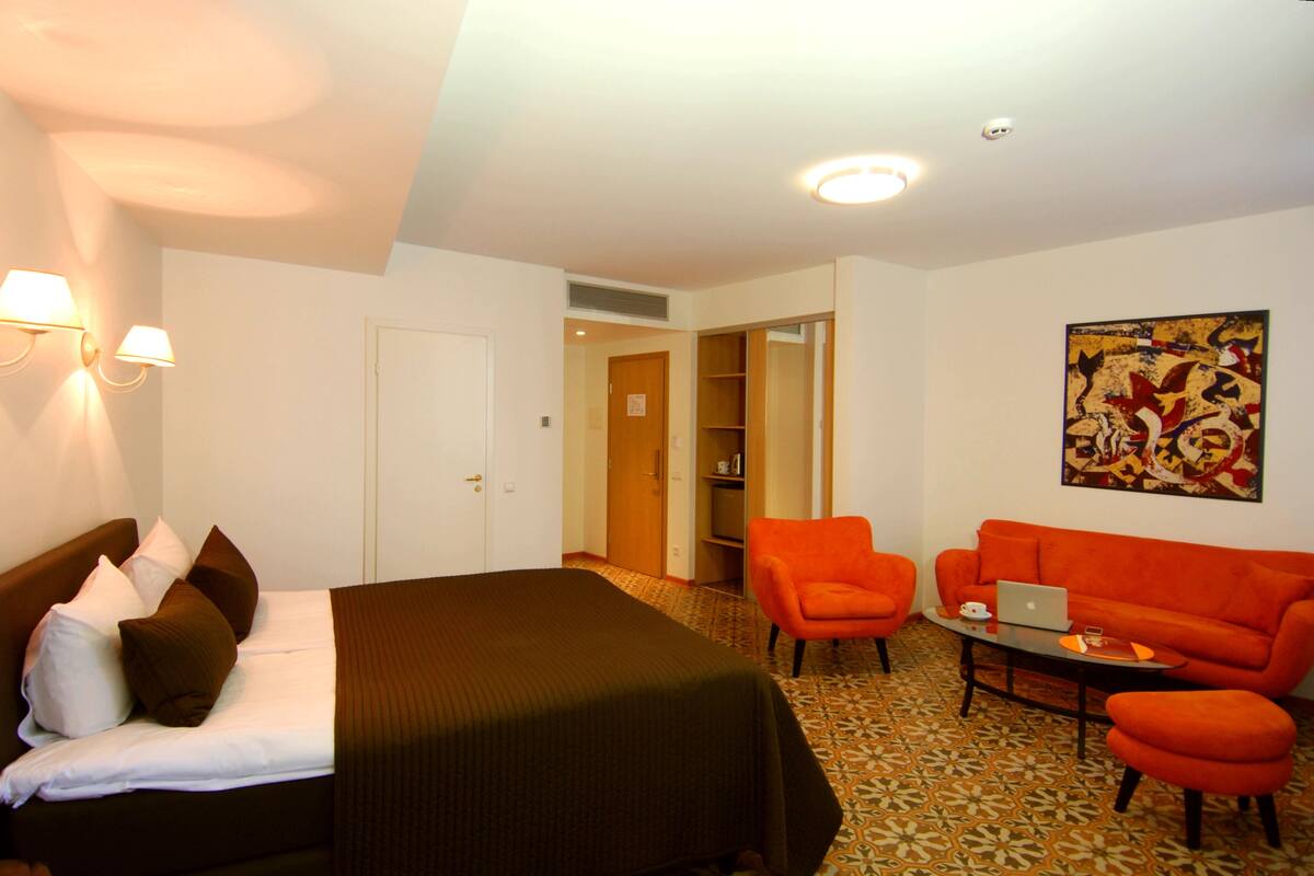 A hotel room with a bed, two orange armchairs, a sofa, a coffee table, and a painting on the wall.