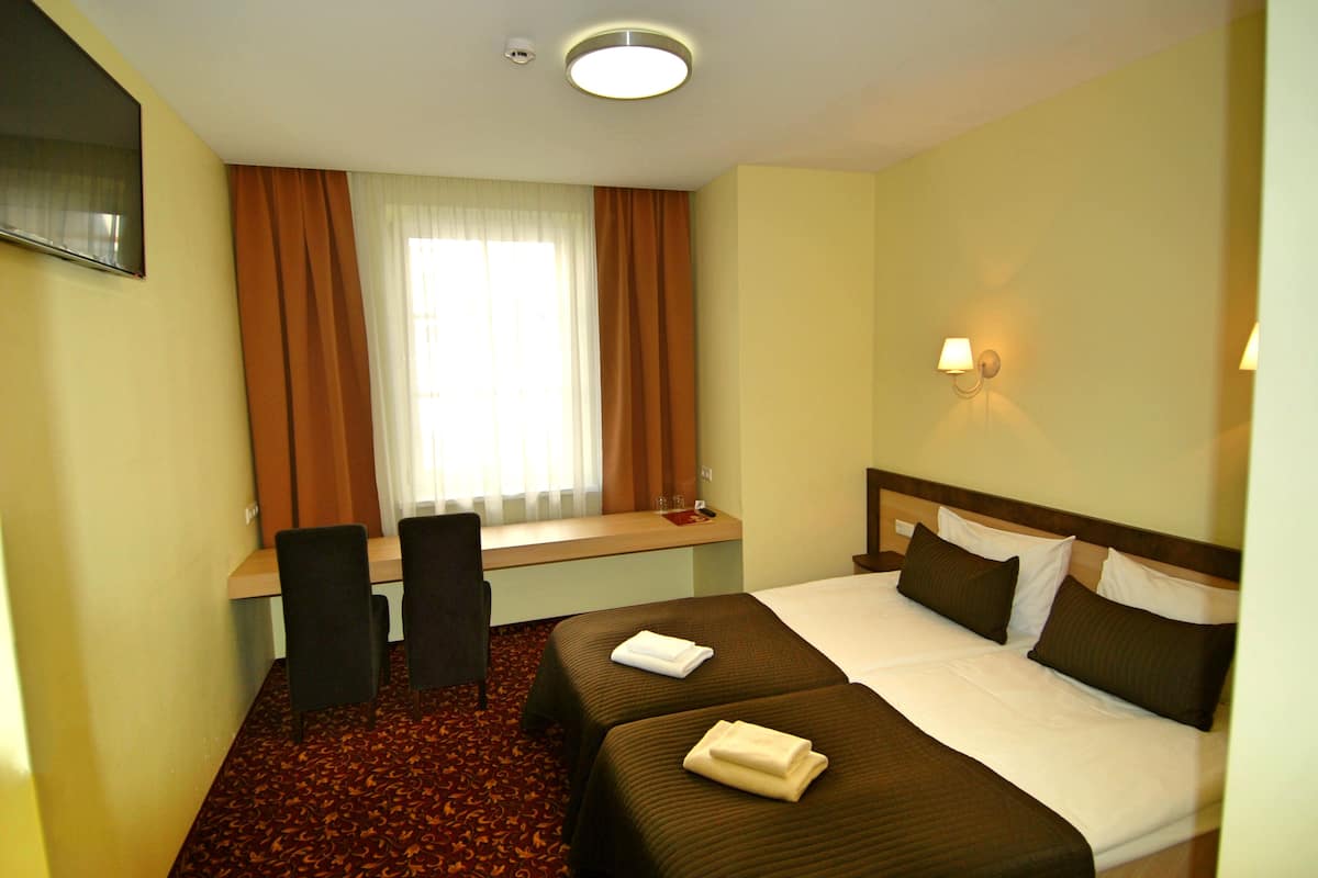 A hotel room with two beds, a desk, a chair, a window with curtains, and a wall-mounted lamp.