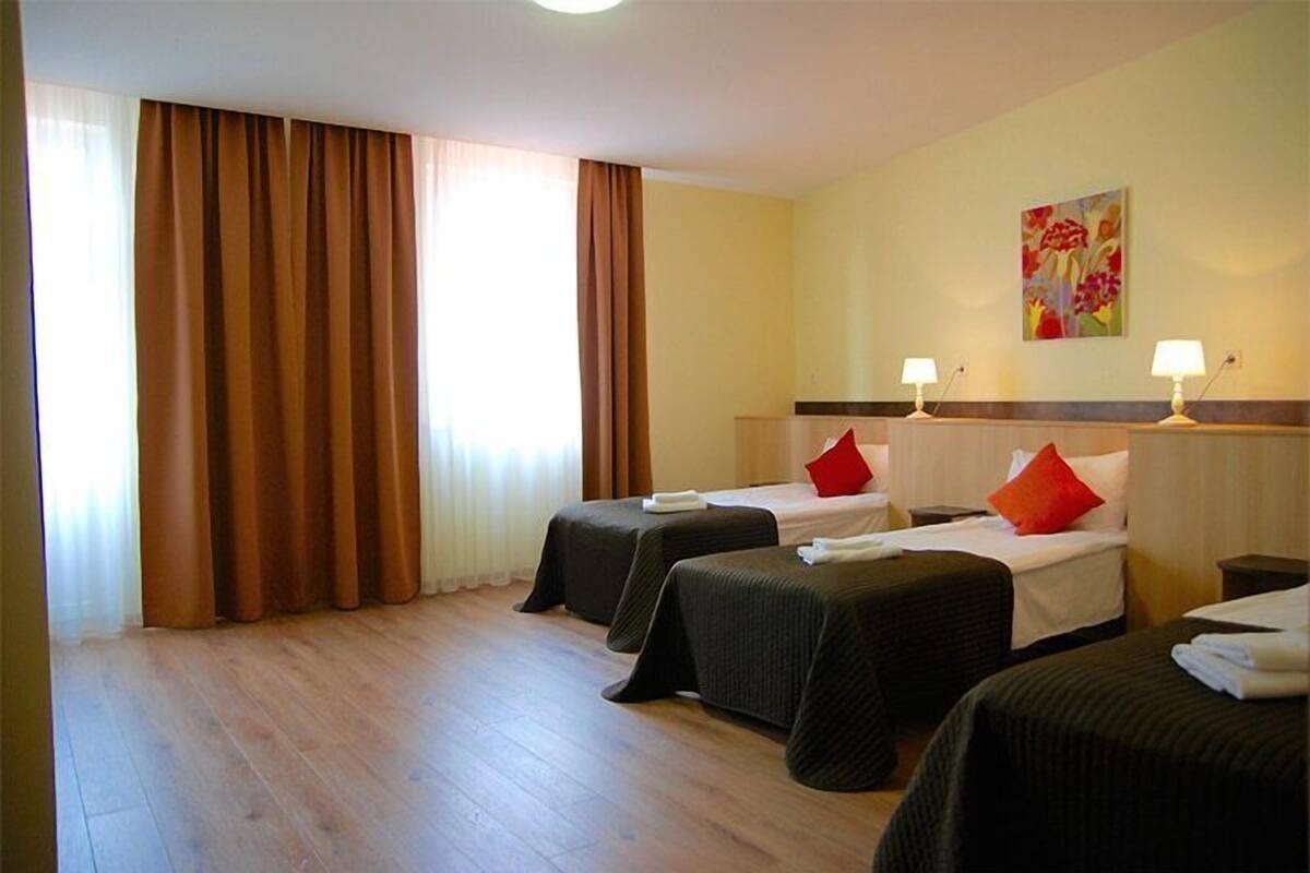 A hotel room with two single beds, a large window with curtains, a painting on the wall, and a nightstand with a lamp.