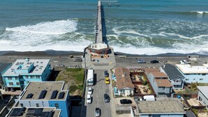 Vista aérea de uma área costeira com um cais que se estende para o mar, edifícios ao longo da costa e uma estrada que leva ao cais.