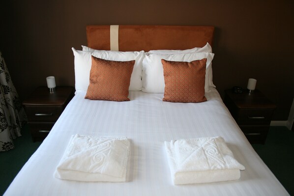 Una cama bien hecha con sábanas blancas y dos almohadones decorativos, una mesita de noche con lámpara, y una cortina parcialmente visible.