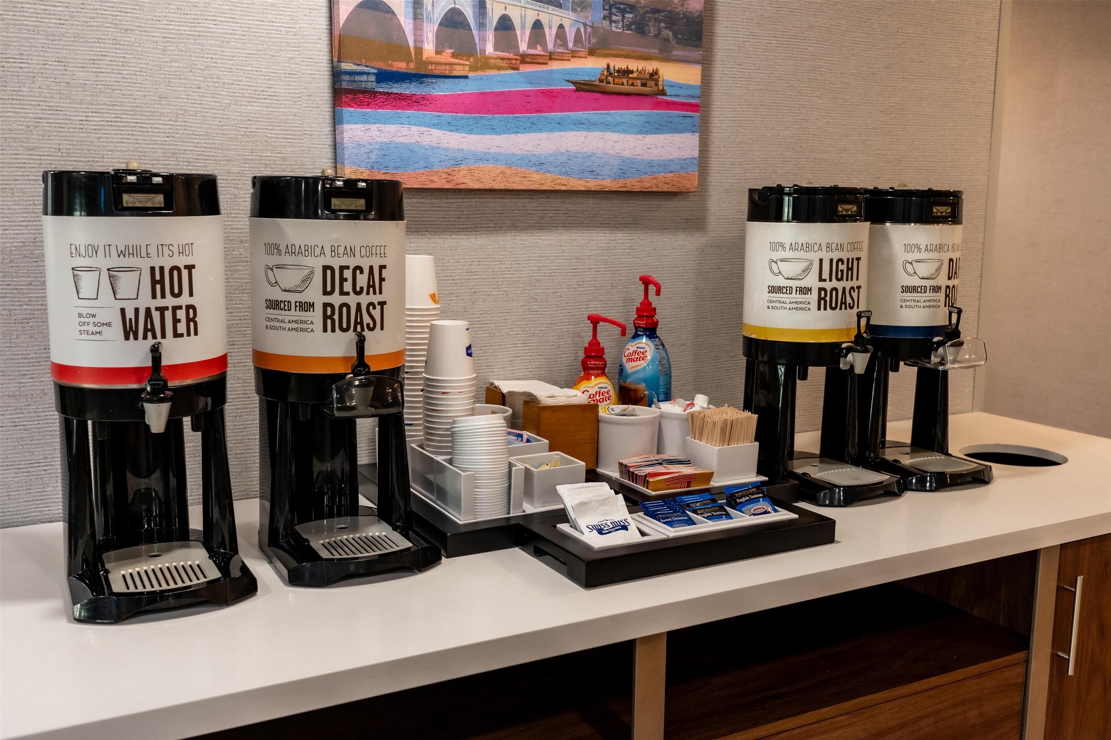 Una estación de café con cuatro dispensadores de café etiquetados como 'Agua Caliente', 'Descafeinado', 'Tueste Suave' y 'Tueste Oscuro'. Hay pilas de tazas blancas, una cafetera y una bandeja con sobres de café y una botella de leche para café.