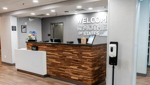 Uma receção com design em madeira, um letreiro com a inscrição 'Welcome to the Mothere of States', um ecrã digital e um dispensador de gel desinfetante.
