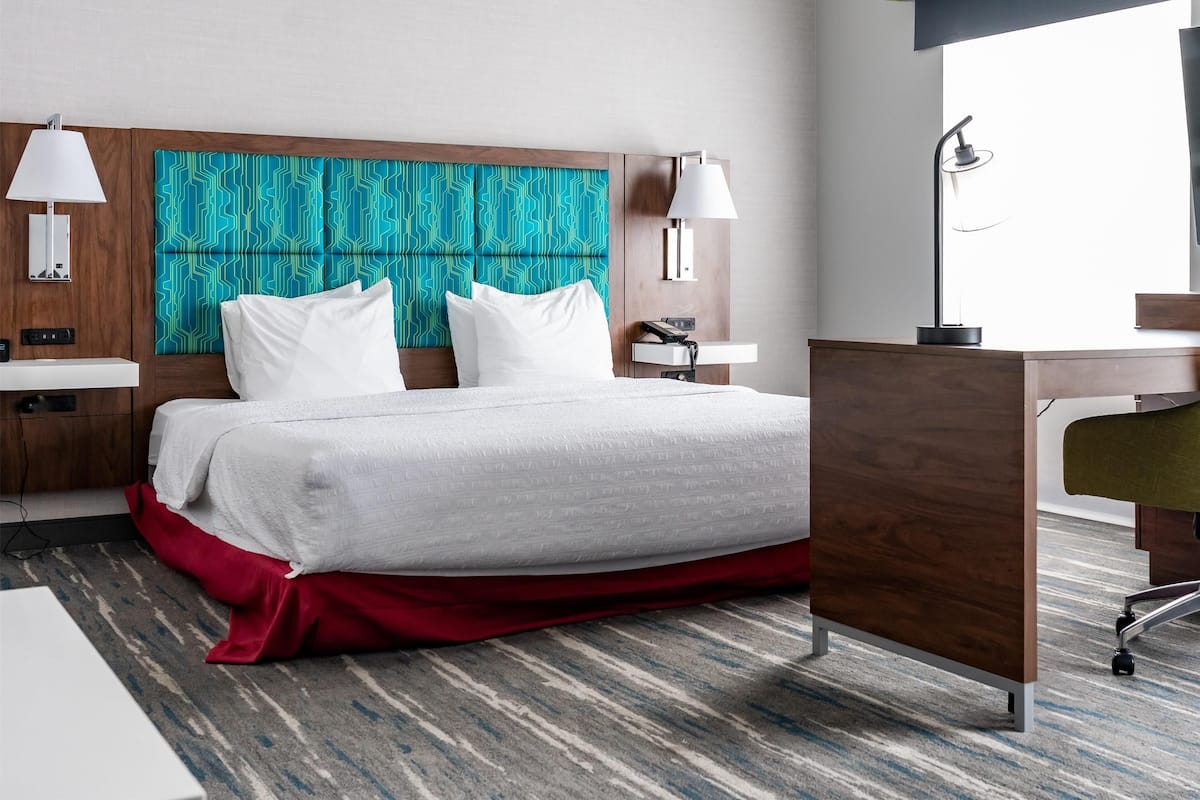 A hotel room with a large bed, two bedside tables, a desk, and a window with blinds.