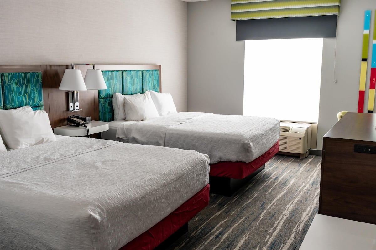 A hotel room with two beds, a nightstand with lamps, a desk, and a window with a view.