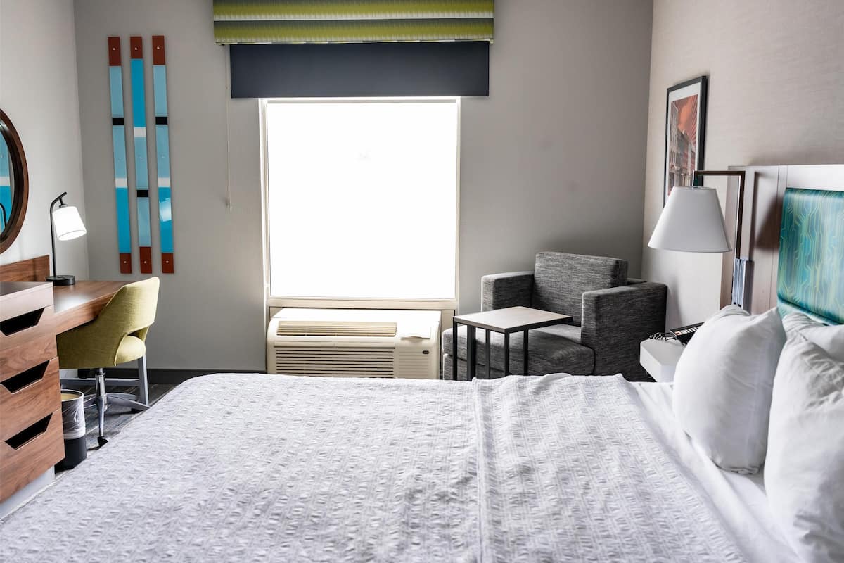 A hotel room with a bed, a desk, a chair, a small table, a lamp, and a window with blinds.