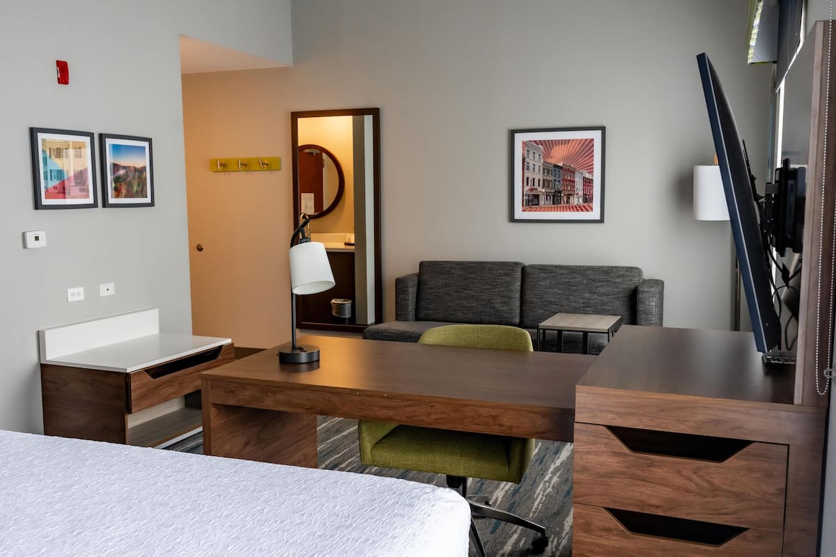 A hotel room with a bed, a desk, a sofa, and a TV. There are framed pictures on the wall and a lamp on the desk.