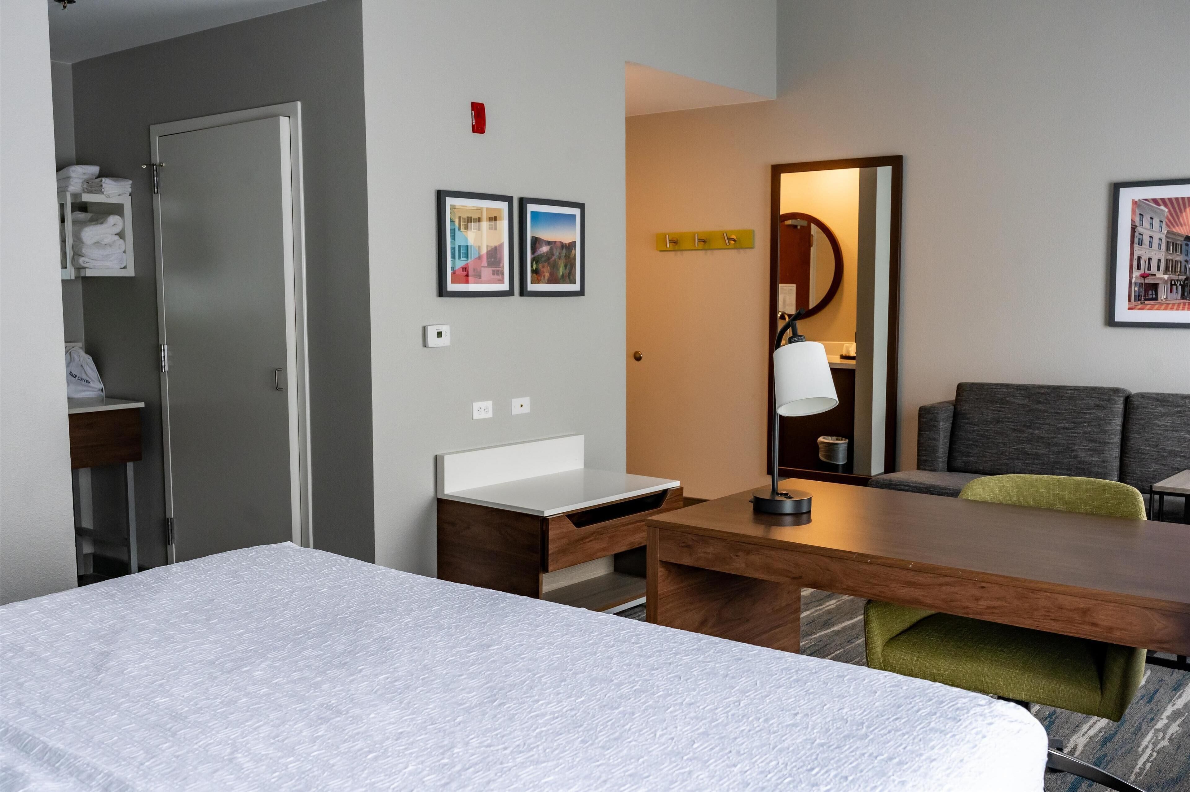 Habitación de hotel con cama, escritorio, sillón, espejo y estante en la pared con toallas.