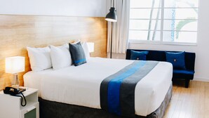 A hotel room with a large bed, a bedside table with a lamp, a nightstand, a blue sofa, and a window with curtains. - Aqua Hotel & Suites (Miami Beach)