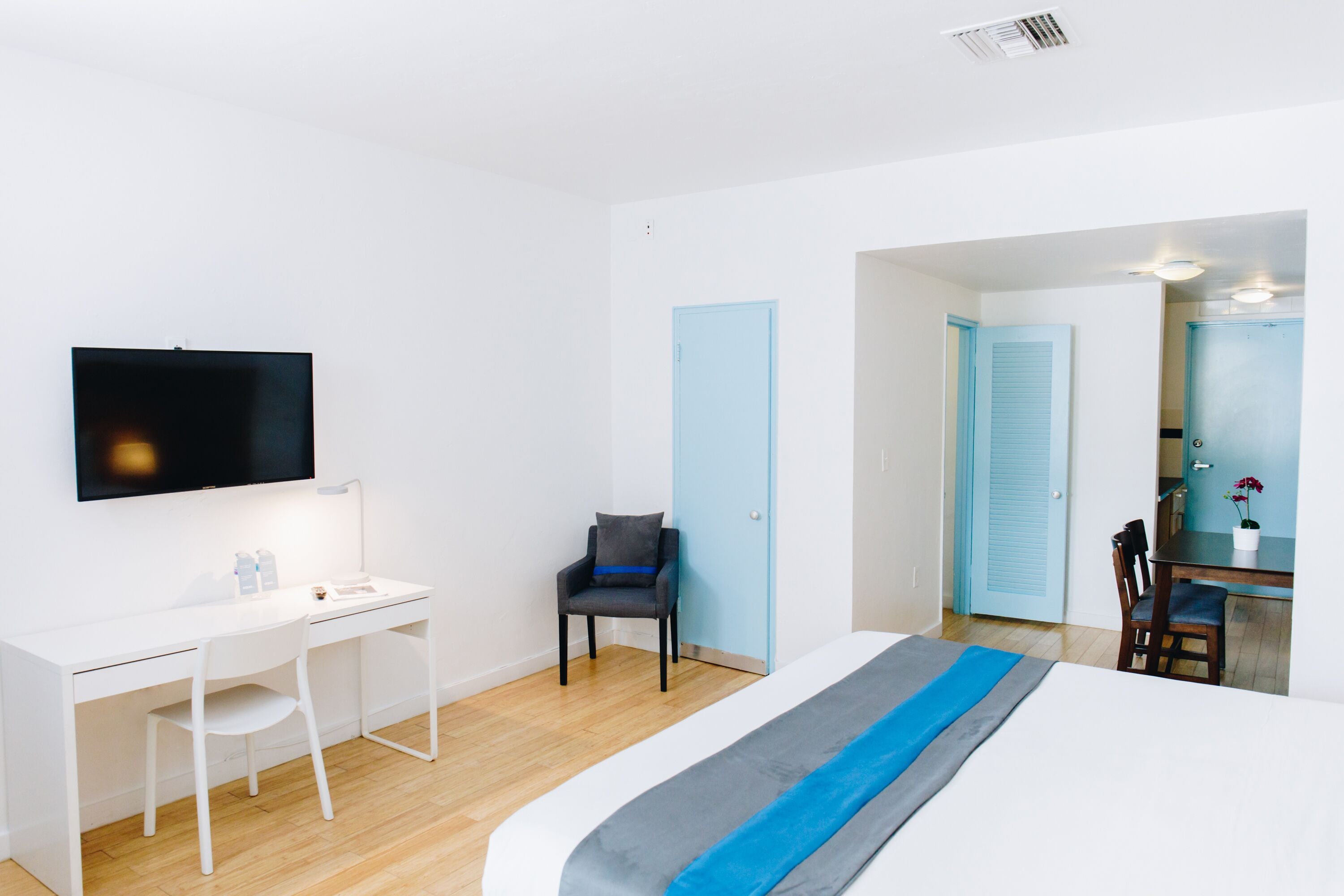 Una habitación de hotel moderna con un escritorio blanco, una cama con cubrecama azul y gris, una silla y un televisor montado en la pared.