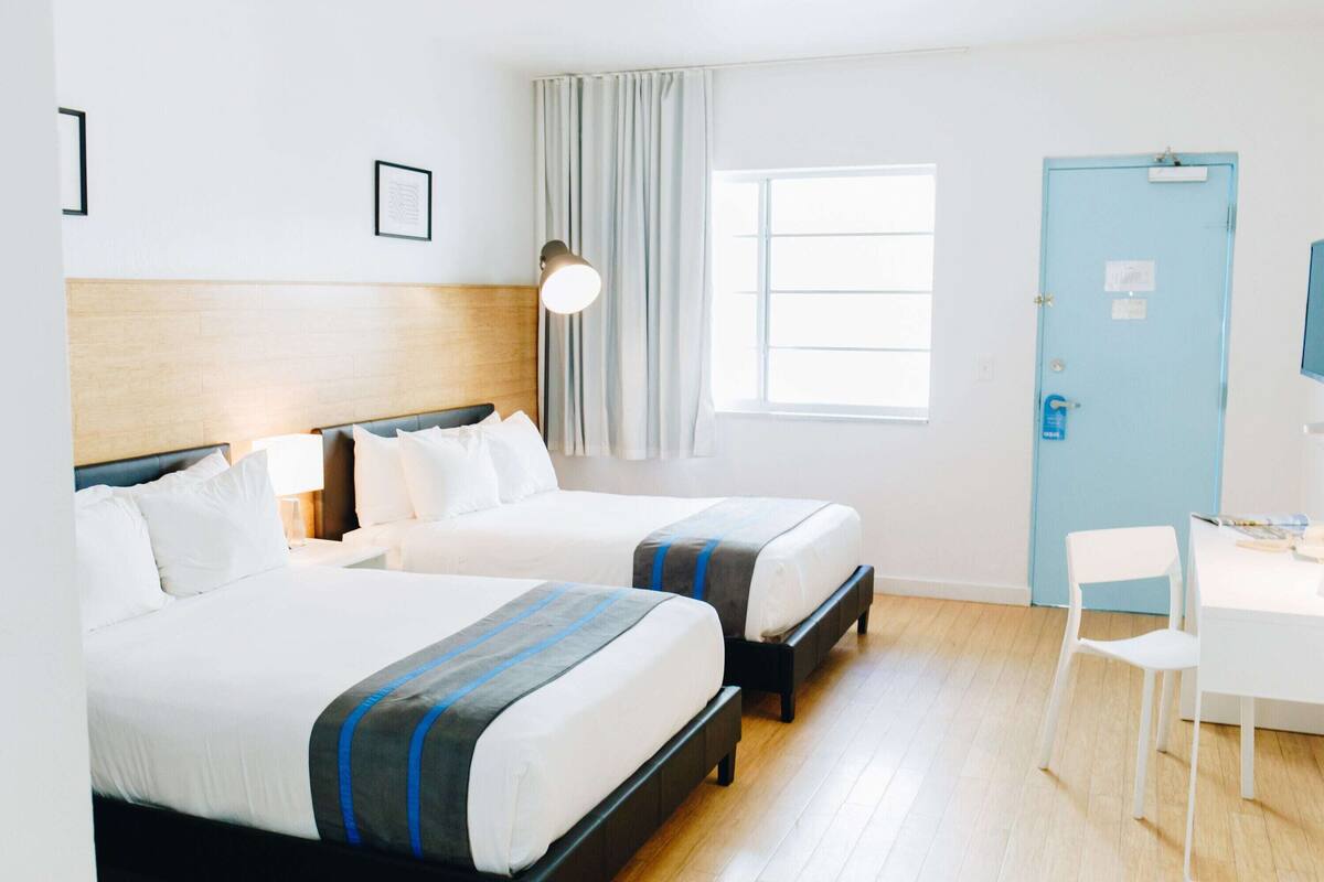 A hotel room with two beds, a desk, a chair, a window, a door, and a lamp.