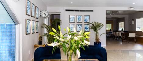 Una sala de estar moderna con un sofá azul, una mesa de centro de vidrio y un florero con flores. Hay cuadros enmarcados en la pared y un reloj.