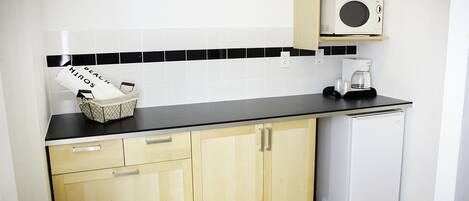 A compact kitchen with wooden cabinets, a black countertop, a microwave, a coffee maker, and a small refrigerator.