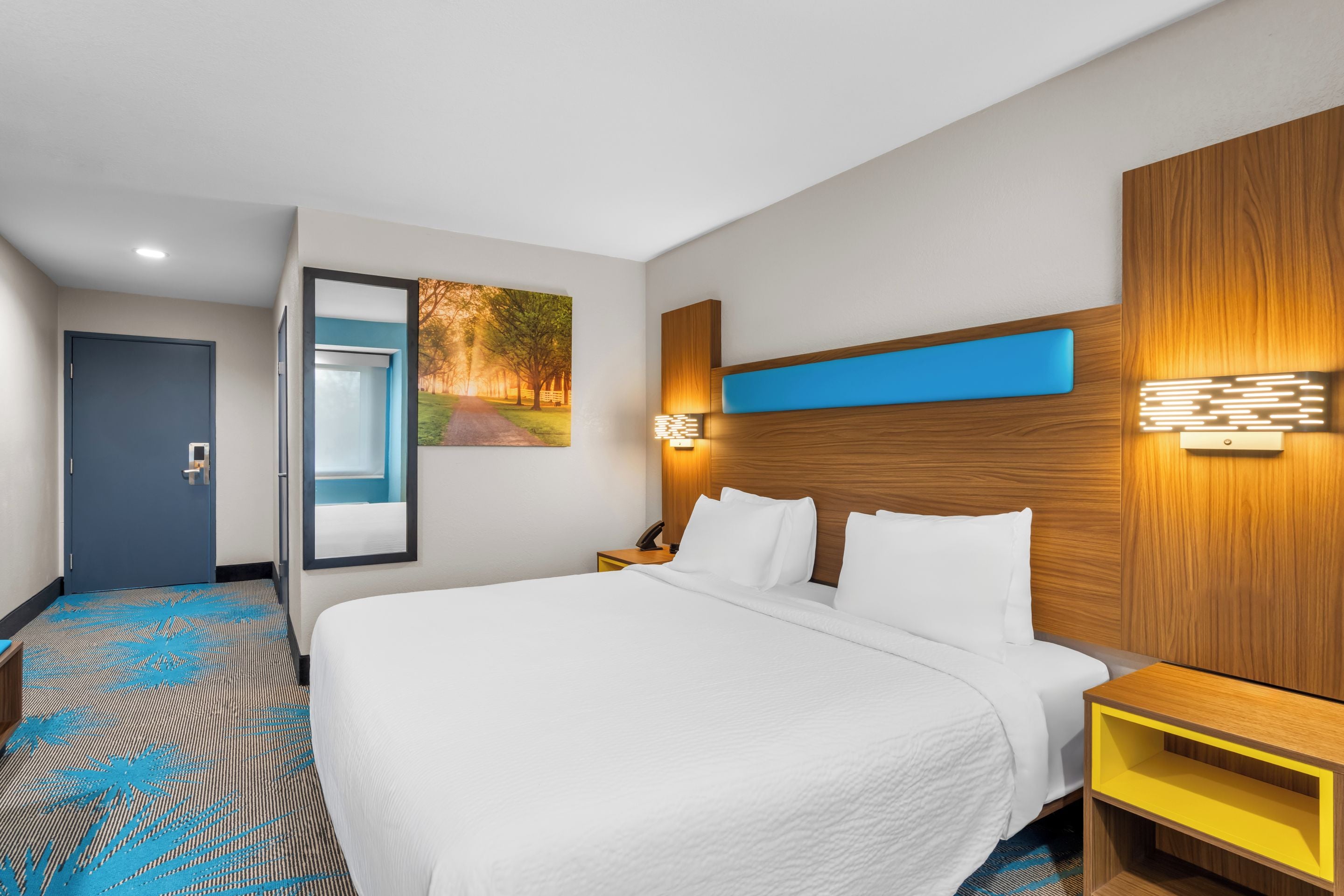 A hotel room with a large bed, a nightstand, a wall-mounted lamp, and a door.