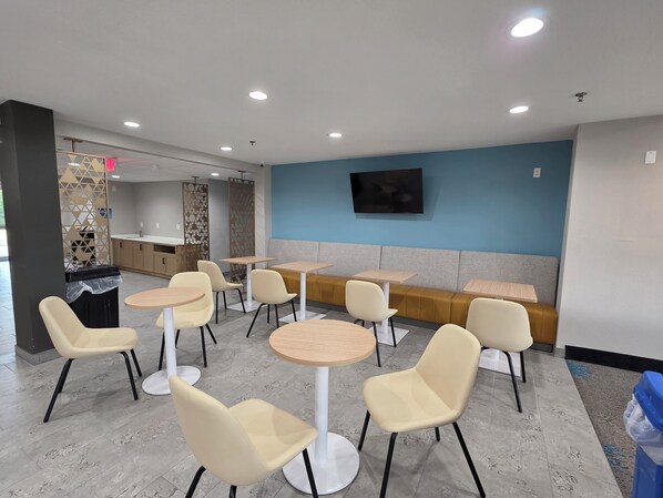 A modern indoor seating area with round tables and cushioned chairs, a flat-screen TV mounted on the wall, and a kitchen area in the background. - Days Inn & Suites by Wyndham Brandon Amphitheater (Brandon)