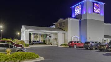 Un Sleep Inn & Suites con un letrero llamativo, un estacionamiento con varios autos y una entrada bien iluminada.